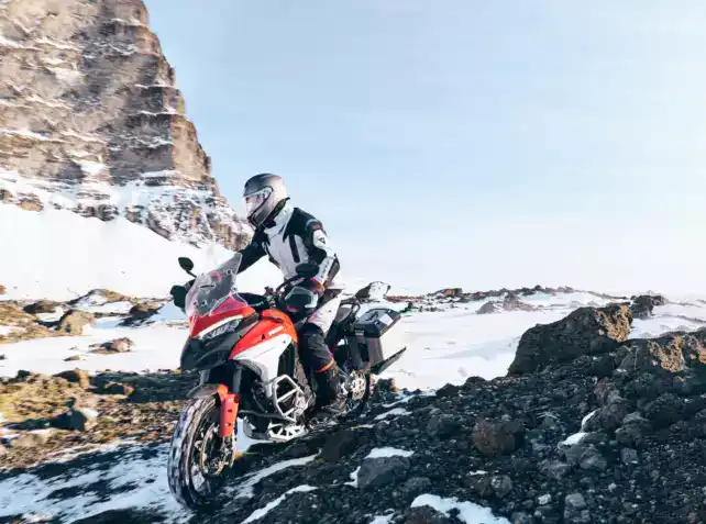 A motorcyclist riding an adventure bike through snowy and rocky mountainous terrain during daytime, wearing full gear including helmet and protective clothing.
