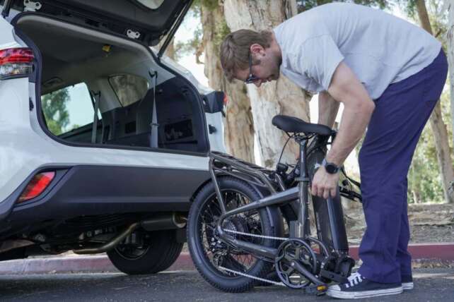 A man preparing an e-bike for riding behind an open car trunk on a sunny day in a park-like setting.