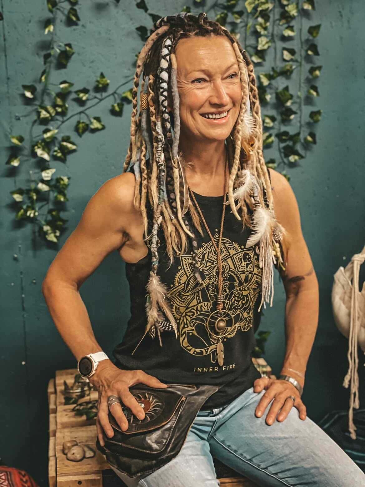 Vibrant woman with dreadlocks smiling, wearing a black graphic tank top and jewelry, standing against a teal wall with green ivy, exuding confidence and free-spirited energy.
